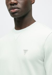 Világoszöld pulóver strukturált kivitelben, bal oldalon háromszög alakú szürke logóval, bordázott kerek nyakkivágással.