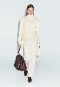 Maglione a collo alto crema, pantaloni bianchi, borsa a tracolla texture marrone scuro e stivali alla caviglia marroni. Modella in piedi contro uno sfondo chiaro.