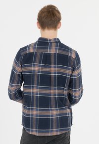 Tummansininen ruudullinen flanellipaita, jossa on vaaleanbeigejä, oransseja ja valkoisia viivoja, varustettuna napitettavalla kauluksella ja selän poimutustyylillä.