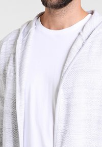 Grå og hvide teksturerede cardigan med åben front, lagdelt over en helt hvid T-shirt, der har et blødt stof og en afslappet pasform.