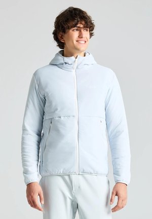 Un joven sonriente luce una chaqueta azul claro con capucha y cremallera, junto con unos pantalones a juego, sobre un fondo liso y claro.