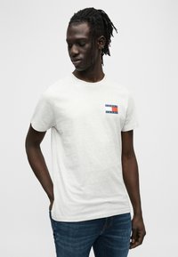 Ελαφρώς γκρι βαμβακερό t-shirt με κοντά μανίκια, που διαθέτει πολύχρωμη ταμπέλα με λογότυπο στο αριστερό στήθος. Φορεμένο με μπλε τζιν παντελόνι.