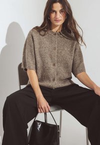 Brūns adīts kardigans ar īsām piedurknēm un apaļu apkakli, ar trim pogām, apvienots ar melnām platām biksēm un mazu melnu somiņu.