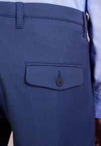 Zbliżenie na ciemnoniebieskie, strukturalne spodnie z kieszenią pod flapą i guzikiem, noszone z jasnoniebieską koszulą.