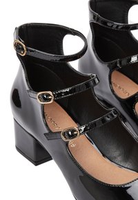 Chaussures en cuir verni noir avec deux sangles de cheville réglables ornées de boucles dorées, un talon carré et un design à bout arrondi.