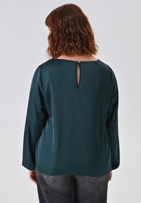 Blusa in satin verde scuro con dettaglio a occhiello sul retro, maniche lunghe e una texture liscia, abbinata a jeans grigio scuro.