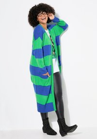 Cardigan lungo a righe verdi e blu con texture a costine, tasche laterali, indossato sopra una t-shirt bianca con grafica e leggings neri, abbinato a stivaletti con borchie.