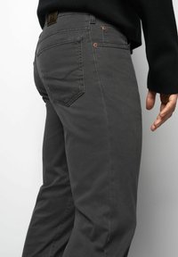 Close-up van een persoon die donkere grijze broek draagt met zichtbare achterzak en koperen klinknagels, zwarte trui-mouw, en de rechterhand naast hun bovenbeen.
