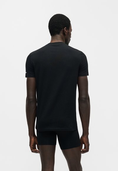 Άνδρας που στέκεται με την πλάτη, φοράει απλό μαύρο κοντομάνικο μπλουζάκι και μαύρα σορτς σε απλό φωτεινό φόντο.