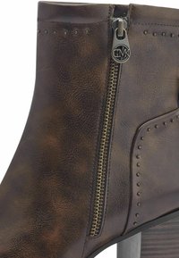 Botín de piel marrón con cremallera lateral, que presenta detalles de tachuelas y un tacón robusto. Acabado texturizado y diseño estilizado.
