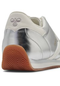 Tylny widok srebrnego trampka z białymi skórzanymi akcentami, haftowanym logo na pięcie oraz brązową gumową podeszwą z wzorem chevron.