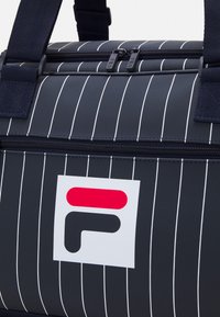 Crni torba sa trakama u bijeloj boji, s dvostrukim crnim patentnim zatvaračem i pravokutnim logom u bijeloj, crvenoj i crnoj boji. Izrađena od izdržljivog materijala.