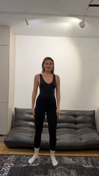 Młoda kobieta w czarnej, bezrękawnikowej bluzce i legginsach stoi boso na wzorzystym dywanie przed ciemnoszarą skórzaną sofą w minimalistycznym pokoju.