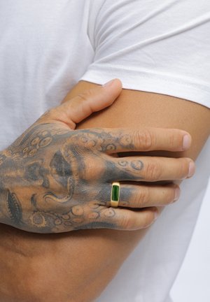 Eine tätowierte Hand mit einem goldenen Ring mit grünem Stein ruht auf dem Arm einer Person, die ein weißes kurzärmeliges Hemd trägt.