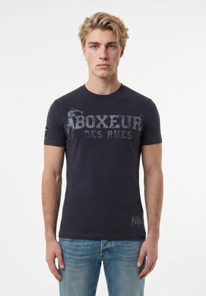 Junger Mann mit blondem Haar, der ein dunkelblaues "Boxeur Des Rues" T-Shirt und hellblaue Jeans vor einem einfarbigen weißen Hintergrund trägt.