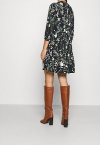 Mujer vistiendo botas de cuero marrón hasta la rodilla y un vestido negro con flores y mangas tres cuartos, de pie sobre un suelo gris liso.