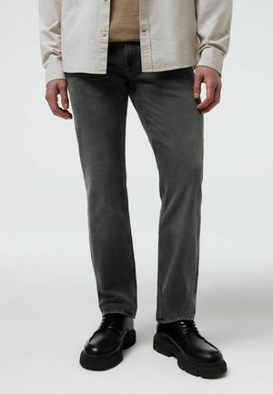 Osoba ubrana w wyblakłe szare dżinsy, beżową koszulę i czarne masywne sznurowane buty stojąca na jasnym tle.