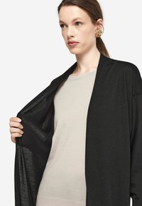 Czarny kardigan otwarty z przodu, wykonany z lekkiego materiału dzianinowego, noszony na beżowej bluzce, ukazujący luźny, swobodny krój i miękką teksturę.