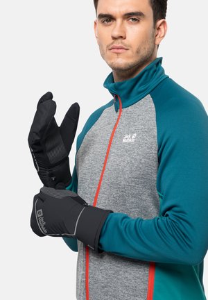 Gants isolés noirs avec des doigts texturés et une prise douce, portés par une personne en veste zippée grise et bleu-vert avec des accents oranges.