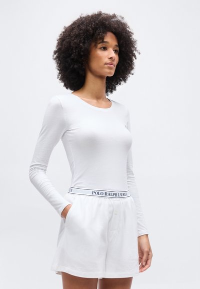 Γυναίκα με σγουρά μαλλιά που φοράει λευκό μακρυμάνικο τοπ και λευκά σορτς με ζώνη Polo Ralph Lauren, στέκεται με το ένα χέρι στην τσέπη.