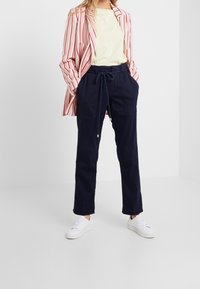 Marinblå joggerbyxor med dragsko i midjan och sidfickor, i kombination med en ljusgul t-shirt och en rödrandig blazer. Vita sneakers.