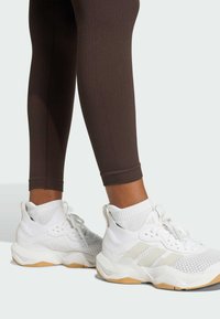 Bílé sportovní boty s texturovanou síťovinou, polstrovanou podrážkou a světle hnědými akcenty; v kombinaci s hnědými žebrovanými legínami.
