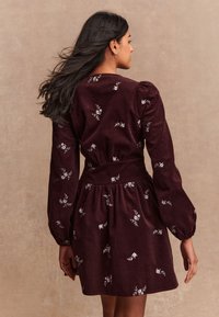 Abito in velluto a coste bordeaux con maniche lunghe a sbuffo, ricamato con motivi floreali bianchi, vita aderente e gonna svasata.