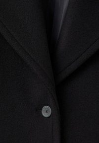 Ingrandimento della parte anteriore di un cappotto di lana nero con un singolo bottone scuro e ampie revers.