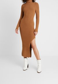 Robe en tricot côtelé marron à manches longues, dotée d'un col montant et d'une fente haute sur la cuisse, associée à des bottines blanches.