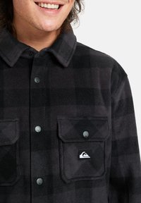 Camisa de franela a cuadros en negro y gris con botones, bolsillos en el pecho y un parche con el logo, que presenta una textura suave y un diseño de cuello clásico.