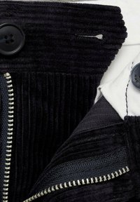 Čierna kordová tkanina s vertikálnymi rebrovaním, ktorá obsahuje hladký zips a detail s gombíkom. Štruktúra zahŕňa kontrastnú bielu podšívku.