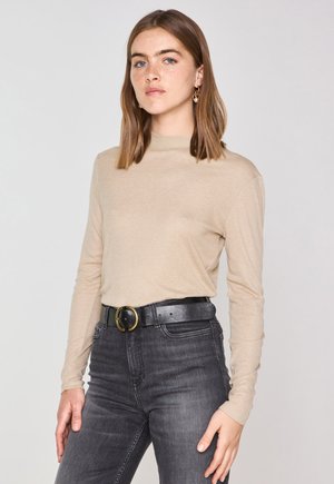 Camicia beige a maniche lunghe con collo alto, indossata con jeans in denim scuro a vita alta e una cintura nera con fibbia dorata.