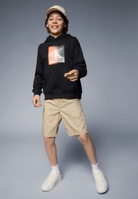 Felpa nera con design grafico, pantaloni beige e sneakers bianchi. La felpa ha una tasca a marsupio e polsini a coste; il cappellino è chiaro con un motivo sottile.