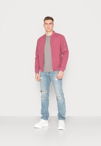 Uma leve jaqueta bomber cor bordeaux, uma t-shirt cinza, jeans claros com efeito desgastado e ténis brancos. Óculos de sol quadrados cor-de-rosa acentuam o visual.