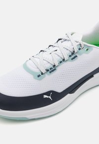 Hvite mesh-sneakers med mørk marineblå aksenter, grønn innvendig og teksturert gummisåle. Har integrert snøresystem og Puma-logo.
