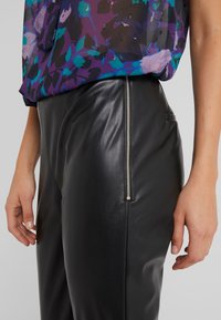 Pantaloni in finta pelle nera con dettaglio della cerniera laterale. Il tessuto è liscio con una leggera finitura lucida. Una parte di un top colorato e con motivo è parzialmente visibile.