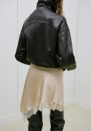 Persoană purtând o jachetă neagră de piele, fustă bej cu dantelă și cizme negre peste genunchi, stând sprijinită de un perete alb cu panouri.