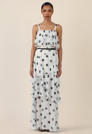 Frau in einem ärmellosen weißen Kleid mit großen schwarzen Polka-Dots und gestuften Rüschen, stehend vor einem schlichten beigen Hintergrund.