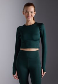 Mørkegrønn atletisk crop top med lange ermer, som har rund hals og glatt, elastisk stoff. Matchet med høytlivsleggings i samme farge.