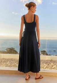 Frau mit einem Dutt, die ein ärmelloses schwarzes Maxikleid und Sandalen trägt, steht auf einer Terrasse mit Blick auf das Meer bei Sonnenuntergang.