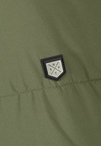 Olivenfarvet stof med en glat tekstur har et sort og sølv hexagonalt logo-patch med præget tekst og et krydsdesign.
