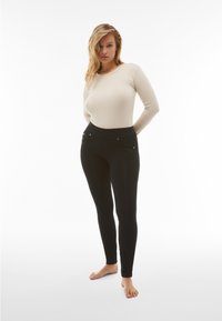Leggings neri con tasche frontali e dettagli in metallo argentato, abbinati a un top aderente a maniche lunghe color crema, in tessuto liscio e leggero.