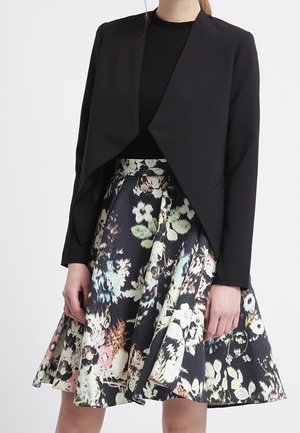 Veste noire cropped avec un col châle, associée à une jupe A-line florale aux couleurs vives et à un motif de tissu texturé.