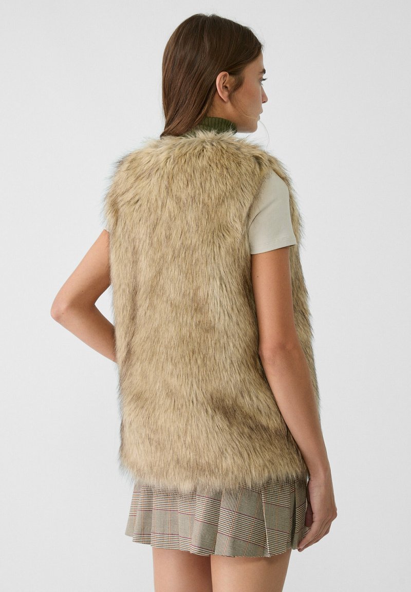 Gilet en fausse fourrure beige avec une texture douce, présentant un design à coupe droite. Porté sur un haut ajusté de couleur claire et associé à une jupe à carreaux.