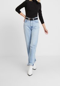 Donna che indossa jeans a vita alta azzurri chiari con risvolti, dolcevita nera, cintura nera e stivaletti bianchi contro uno sfondo bianco.