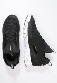 Baskets de sport noires avec tige en maille, lacets élastiques et semelle blanche tachetée. Présente le logo Reebok et un col rembourré pour plus de confort.