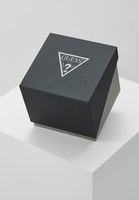 Cutie neagră cu textură, cu un design triunghiular, având un logo argintiu "GUESS?" în partea de sus și o bază gri.