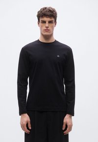 Musta pitkähihainen T-paita, joka on valmistettu puuvillasta pyöreällä pääntiellä ja pienellä pyöreällä logolla vasemmassa rinnassa. Slim fit ja sileä tekstuuri.