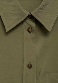 Chemise vert olive avec un col classique, une patte de boutonnage à boutons marron et une poche plaquée. Le tissu présente une finition lisse avec une légère texture.
