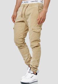 Pantalons cargo beiges avec taille ajustable, poignets élastiques, poches latérales et finition texturée, portés avec des baskets blanches.